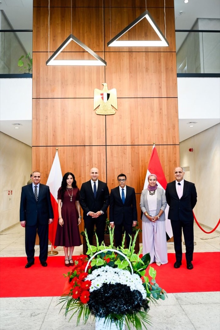 Six guests on a red carpet at the Egyptian Embassy in Warsaw on Egypt National Day, between Polish and Egyptian flags below the Egyptian state emblem.